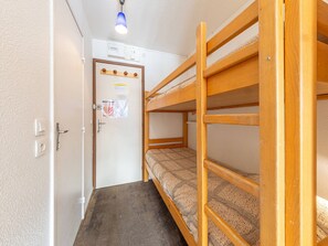 Een smalle gang met een houten stapelbed aan de rechterkant, een deur met lichtschakelaar en een bericht op de muur, en een plafondlamp.