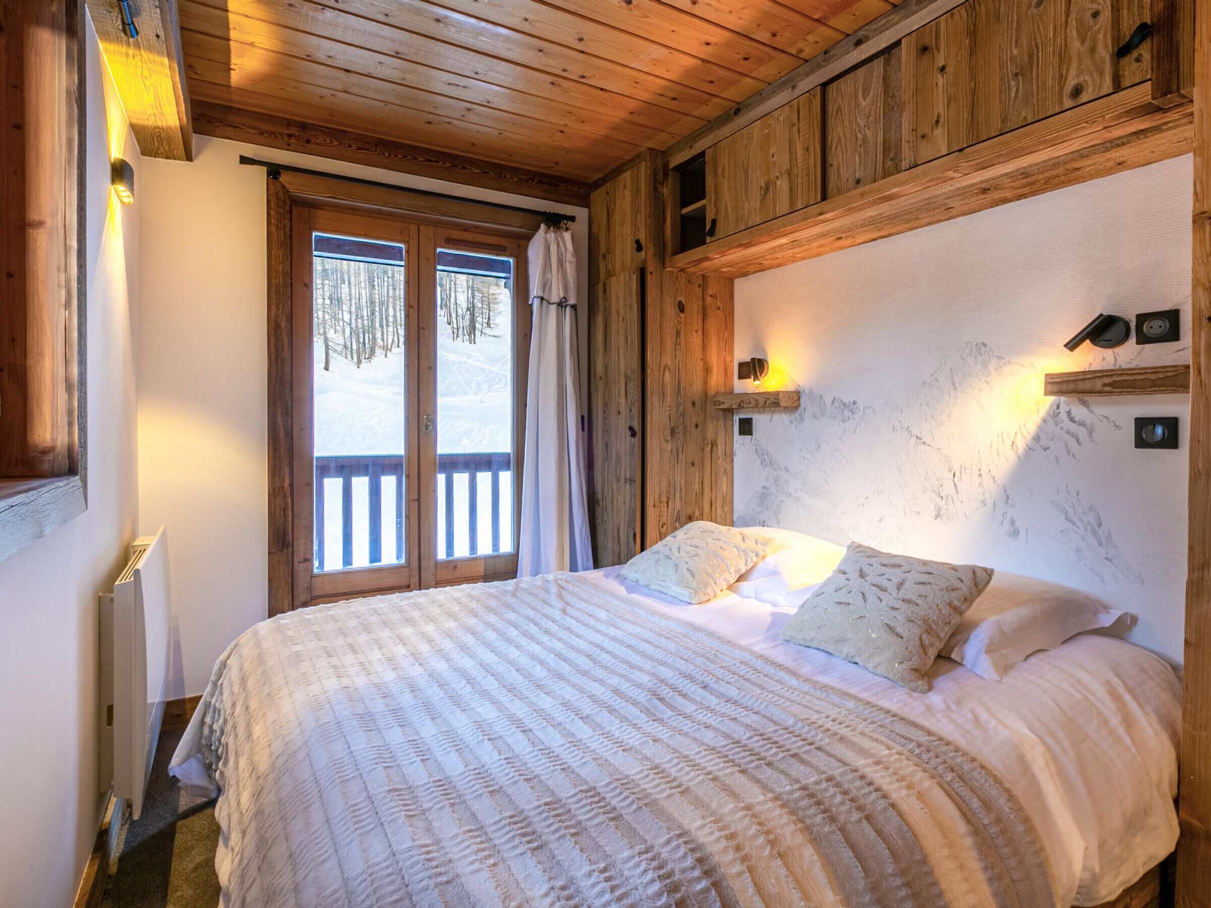 Ein gemütliches Schlafzimmer mit einem großen Bett, Holzbalkendecke und Blick auf eine verschneite Landschaft durch eine Schiebetür.