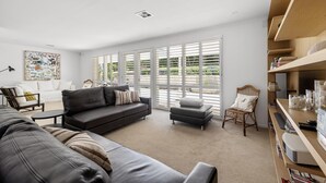 Uma sala de estar moderna com um sofá de couro preto, uma poltrona branca e uma mesa pequena. Janelas amplas com persianas permitem a entrada de luz natural, e uma estante é visível à direita.
