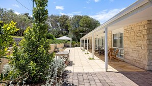 Uma área de varanda coberta com móveis de exterior, parede de pedra e um jardim com diversas plantas.