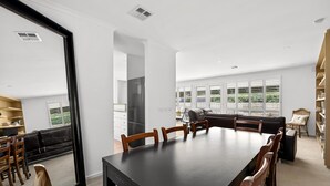 Uma sala de jantar ampla com uma mesa comprida e cadeiras, um sofá preto e vista para o verde através de grandes janelas.