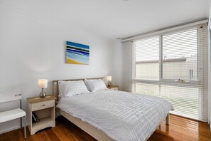 A bedroom with a bed, bedside tables, a nightstand, a window with blinds, and a painting on the wall.