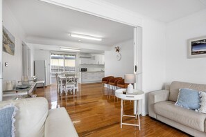 A modern living room with a sofa, a dining area with chairs, a kitchen in the background, and a hallway leading to another room.