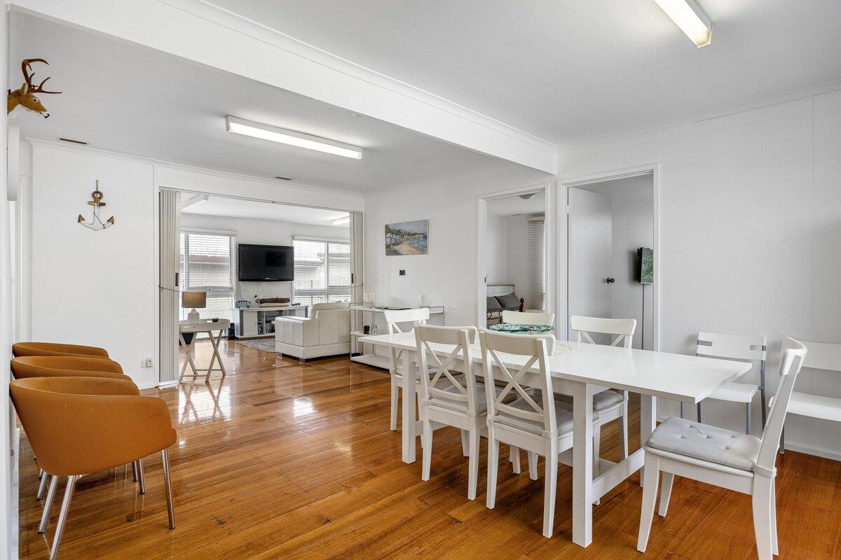 Een ruime woonkamer met een eettafel en stoelen, een keuken op de achtergrond, en een wit kleurenschema.