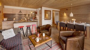 Ein gemütlicher Wohnbereich mit einer Couch, einem Couchtisch und Ledersesseln. Eine Küche mit Holzausstattung und ein Essbereich mit Tisch für vier Personen sind zu sehen.