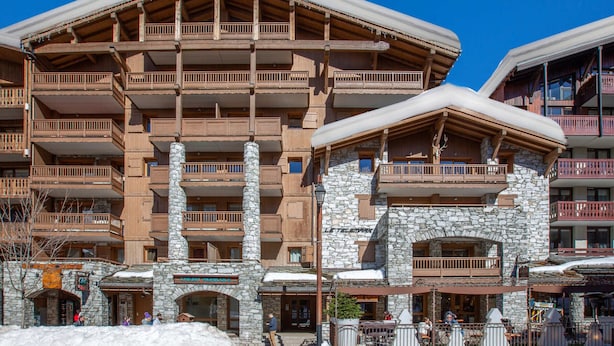 Ein mehrstöckiges Hotel mit Stein- und Holzbauweise, Balkonen und einer Skipiste im Hintergrund.