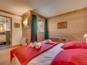 A hotel room with a large bed, a window with curtains, a wooden wall, and a small table with a lamp. - Luxurious 4-Room Apartment with South-Facing Balcony in Central Val-d'Isère (Val-d'Isère)