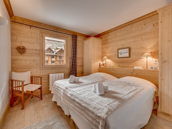 Une pièce en bois avec un lit, une chaise, une fenêtre donnant sur des immeubles, une lampe et un tableau accroché au mur.