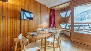 Un intérieur en bois avec une table à manger dressée pour quatre personnes, un téléviseur à écran plat fixé au mur et de grandes fenêtres offrant une vue sur un paysage enneigé.