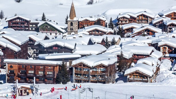 Un village enneigé, avec des bâtiments en bois, un clocher d’église et des pistes de ski.