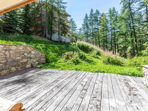 A wooden deck with a stone wall, surrounded by lush greenery and tall trees. - Stunning High-End Mountain Apartment with Refined Charm in Val-d'Isère (Val-d'Isère)