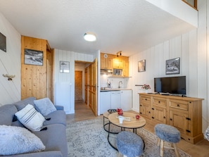 A cozy living room with a sofa, a coffee table, and a TV. There's a kitchen area with wooden cabinets and a dining table.