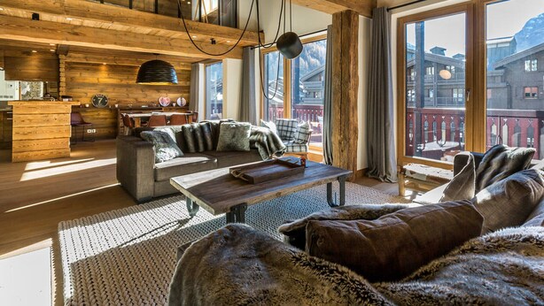 Een ruime woonkamer met een houten plafond, een zithoek, een eettafel en uitzicht op een bergachtig landschap door grote ramen.
