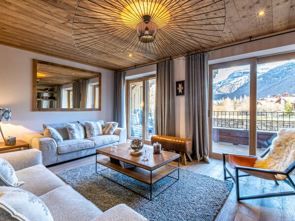 A modern living room with a wooden ceiling, a large mirror, a sofa, a coffee table, and a view of mountains through large windows.