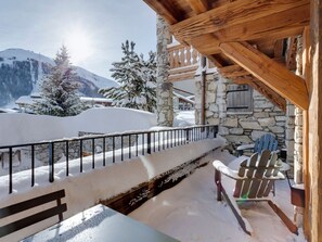 A snow-covered outdoor area with a stone building, wooden beams, a bench, and a chair.