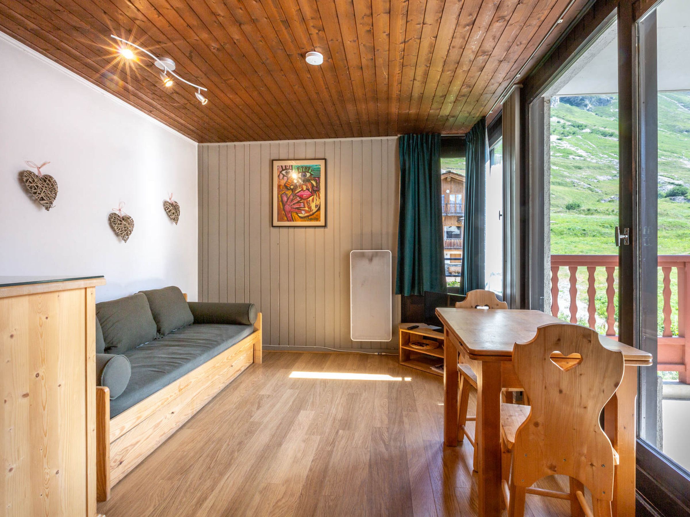 Una stanza accogliente con soffitto in legno, un divano verde, un tavolo in legno e vista su un paesaggio verdeggiante attraverso un'ampia finestra.