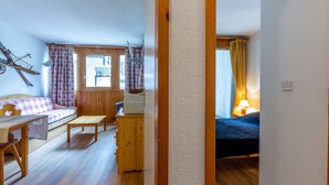 A cozy room with a wooden floor, a bed, a desk, and a chair. There is a window with curtains and a lamp on the desk.