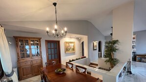 Un espace repas comprenant une table et des chaises en bois, un lustre, un buffet aux portes vitrées et un escalier menant à un autre niveau.