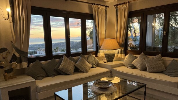 Un salon moderne avec une table basse en verre, des canapés de couleur beige et une vue sur un coucher de soleil se déclinant sur un paysage montagneux.