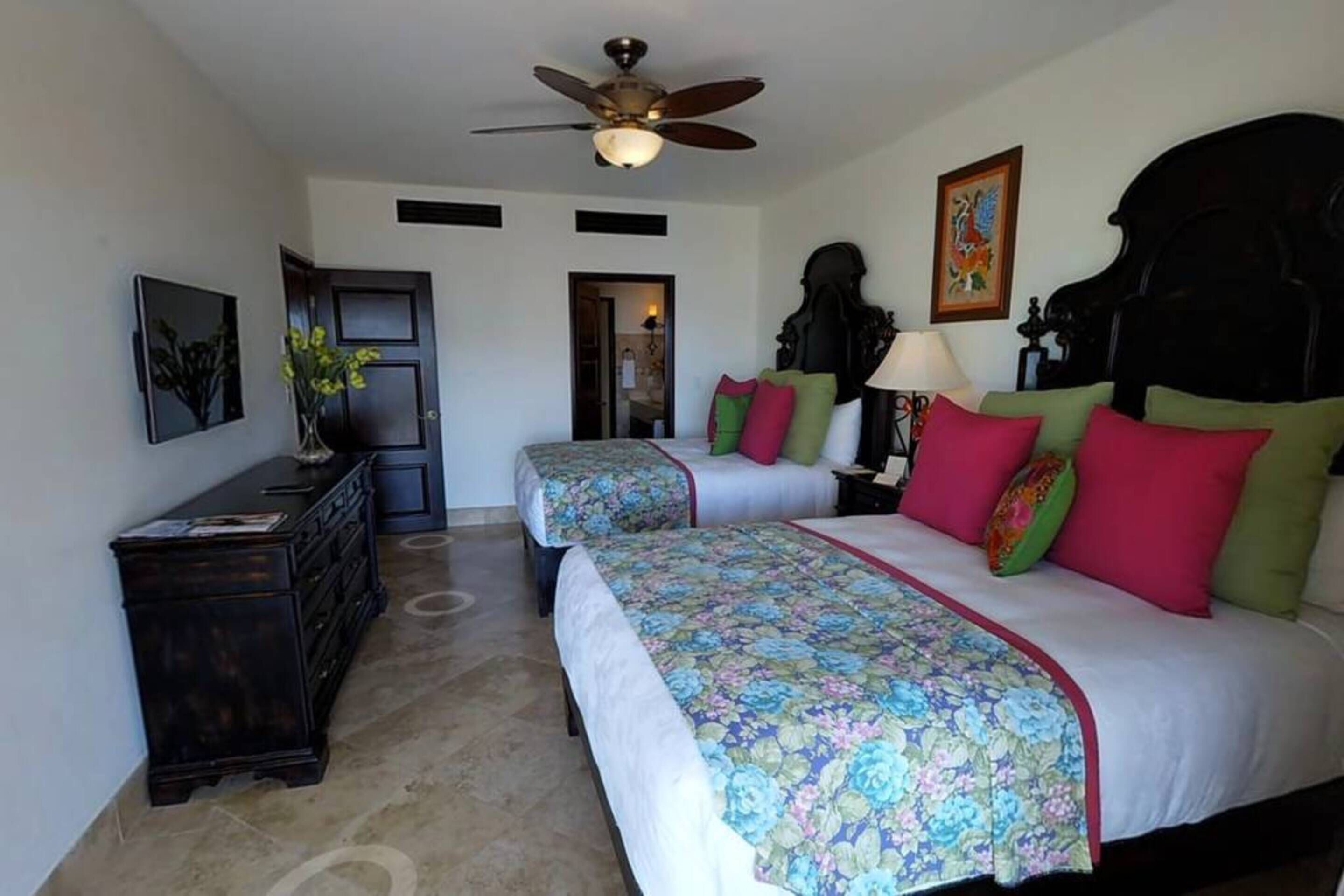 Une chambre d’hôtel avec deux lits, un ventilateur de plafond, une commode et une porte donnant sur une autre pièce.