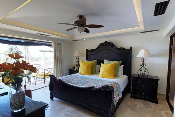 A bedroom with a large bed, two bedside tables, a ceiling fan, and a view of the outdoors.