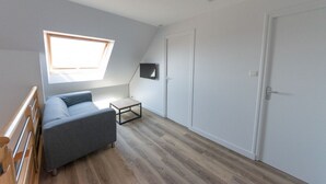 Une pièce avec un plafond en pente, un banc intégré, une petite table, un canapé gris et un téléviseur fixé au mur.