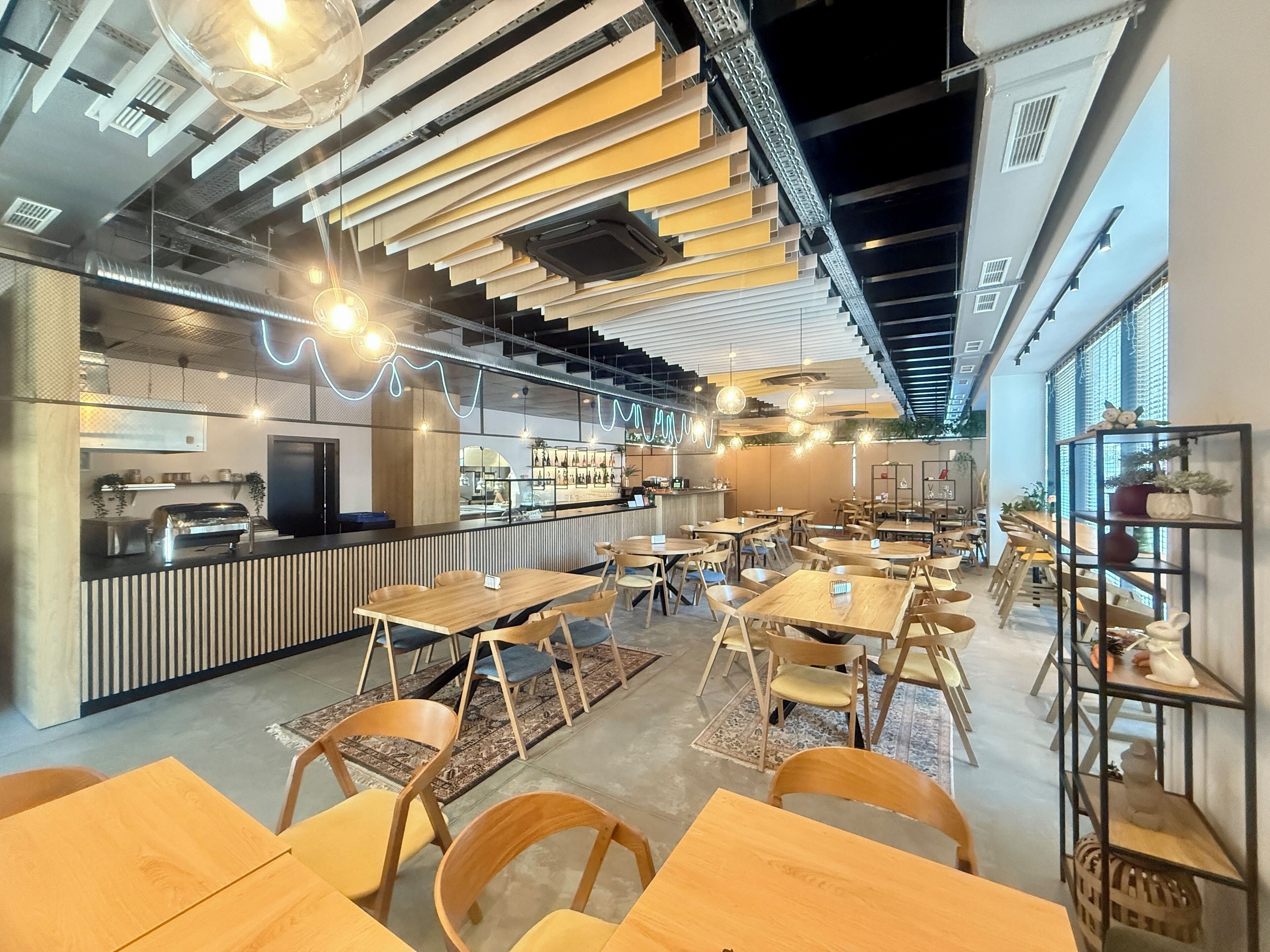 Un restaurante moderno con mesas y sillas de madera, una zona de bar y un techo con vigas a la vista y luces colgantes.