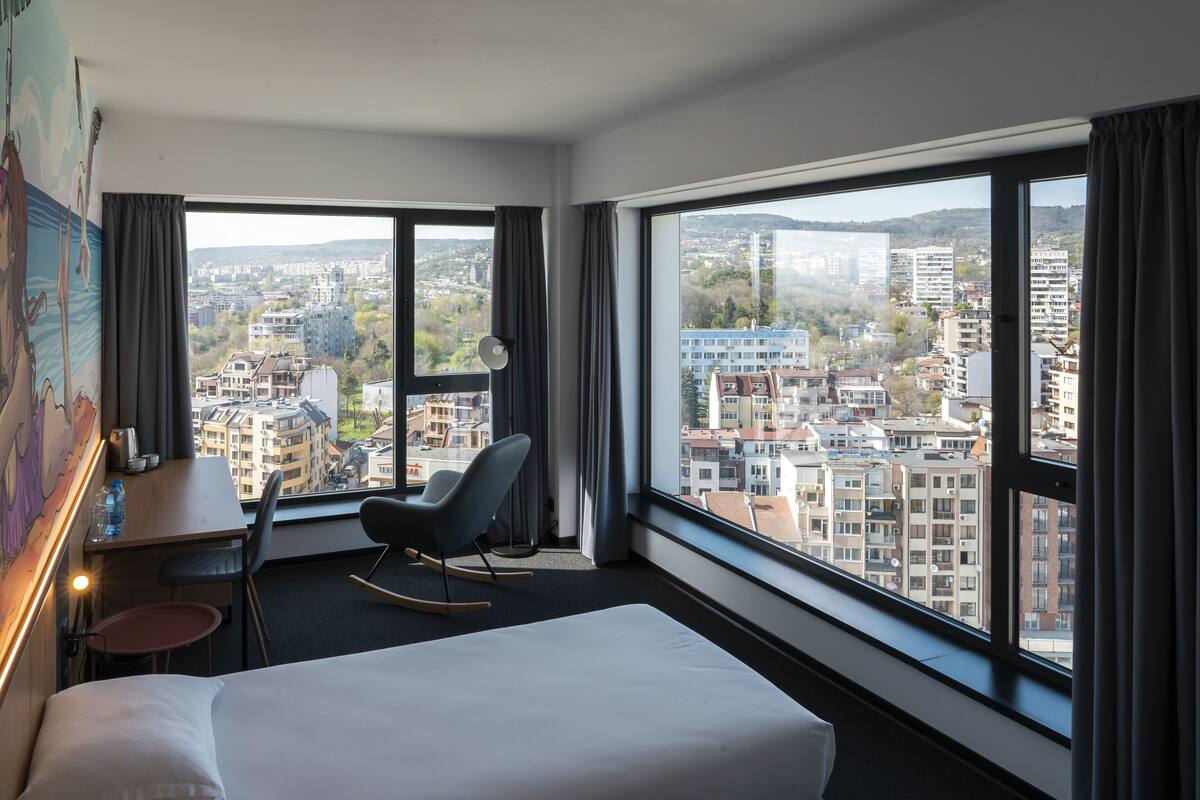 A hotel room with a bed, a desk, a chair, and large windows offering a city view.