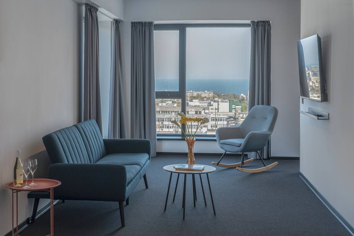 A modern hotel room with a sofa, a rocking chair, a small table with a vase, and a TV on the wall.