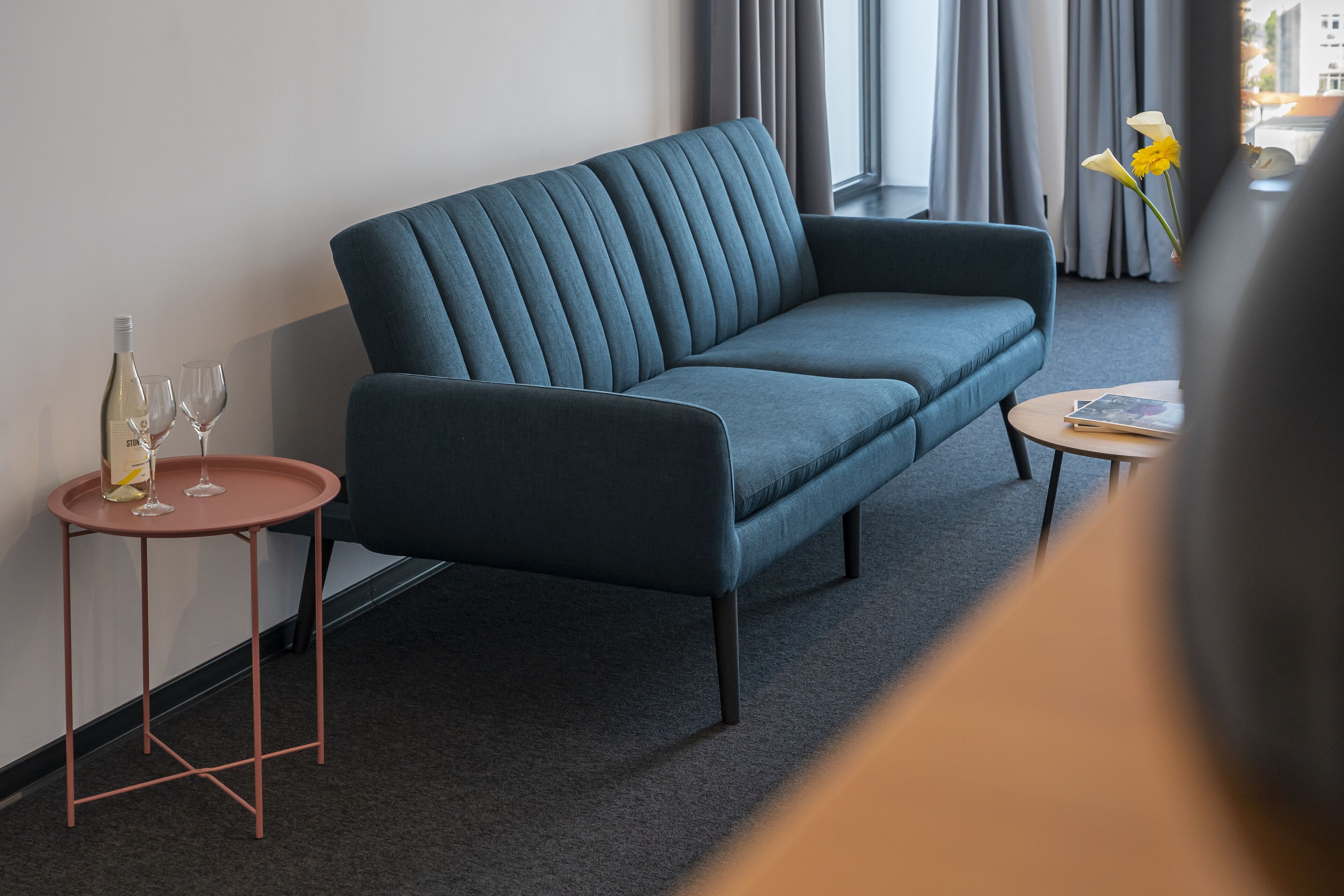 Una sala de estar moderna con un sofá azul oscuro, una mesita redonda con una botella de vino y copas, y un jarrón con flores.