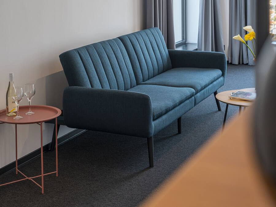Una sala de estar moderna con un sofá azul oscuro, una mesita redonda con una botella de vino y copas, y un jarrón con flores.