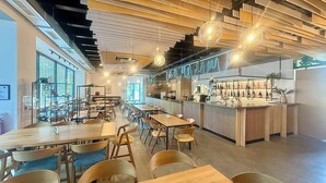 A modern restaurant interior with wooden tables and chairs, a bar area with shelves of bottles, and pendant lighting.