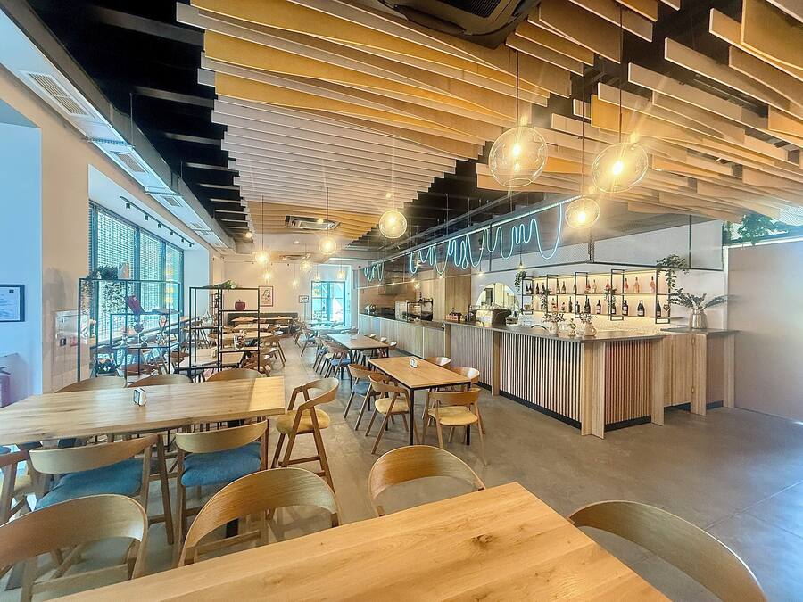 Un interior de restaurante moderno con mesas y sillas de madera, una zona de bar con estantes llenos de botellas e iluminación colgante.