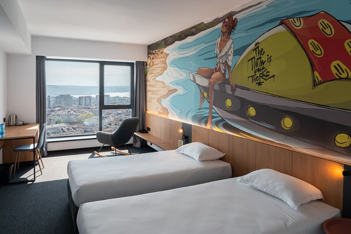 A hotel room with two beds, a desk, a chair, and a large window offering a city view.