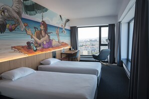 A hotel room with two beds, a desk, a chair, a large window with a city view, and a mural on the wall. - Campus 90 (Varna)