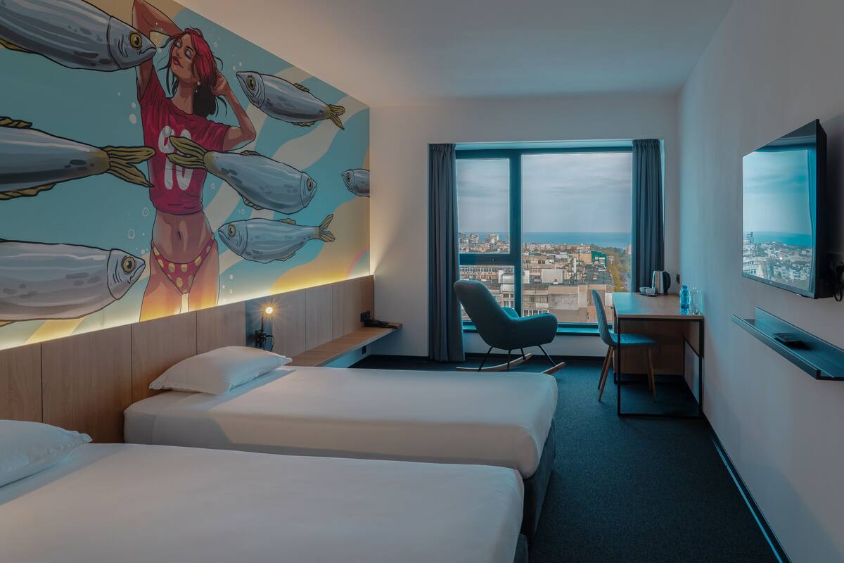 A hotel room with two beds, a desk, a chair, a large window with a city view, and a wall mural.
