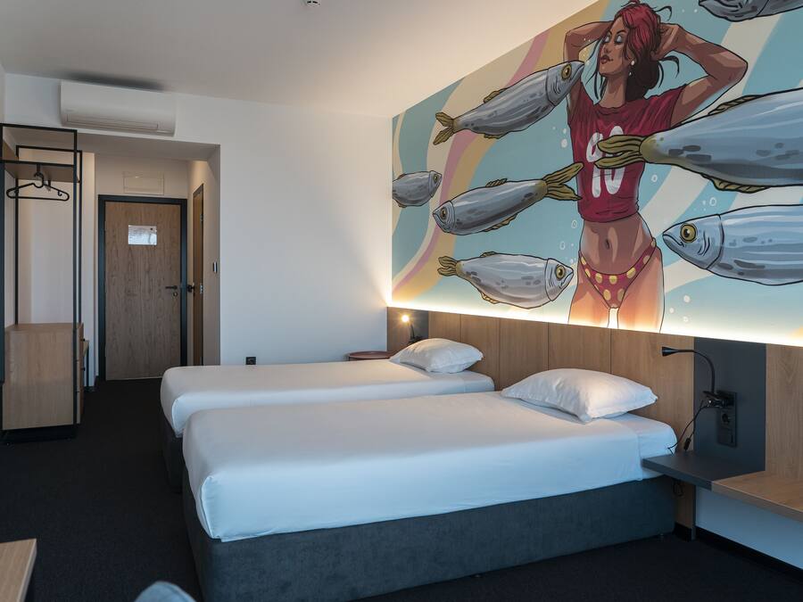 A hotel room with two beds, a wooden wardrobe, a desk, and a wall with a large fish-themed mural.