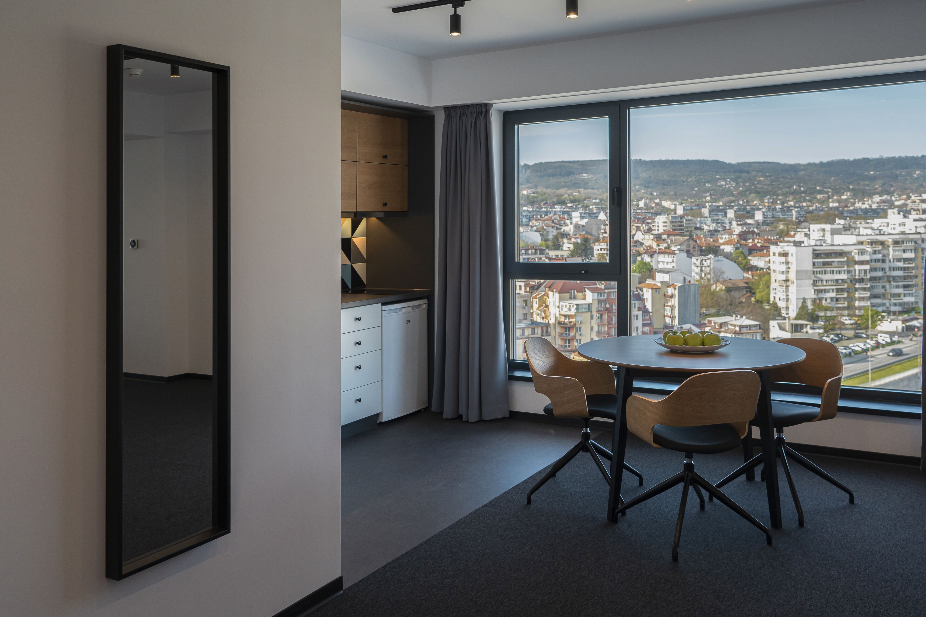 Quarto de hotel moderno com uma janela grande que oferece vista da cidade, uma mesa redonda com cadeiras e uma pequena cozinha.