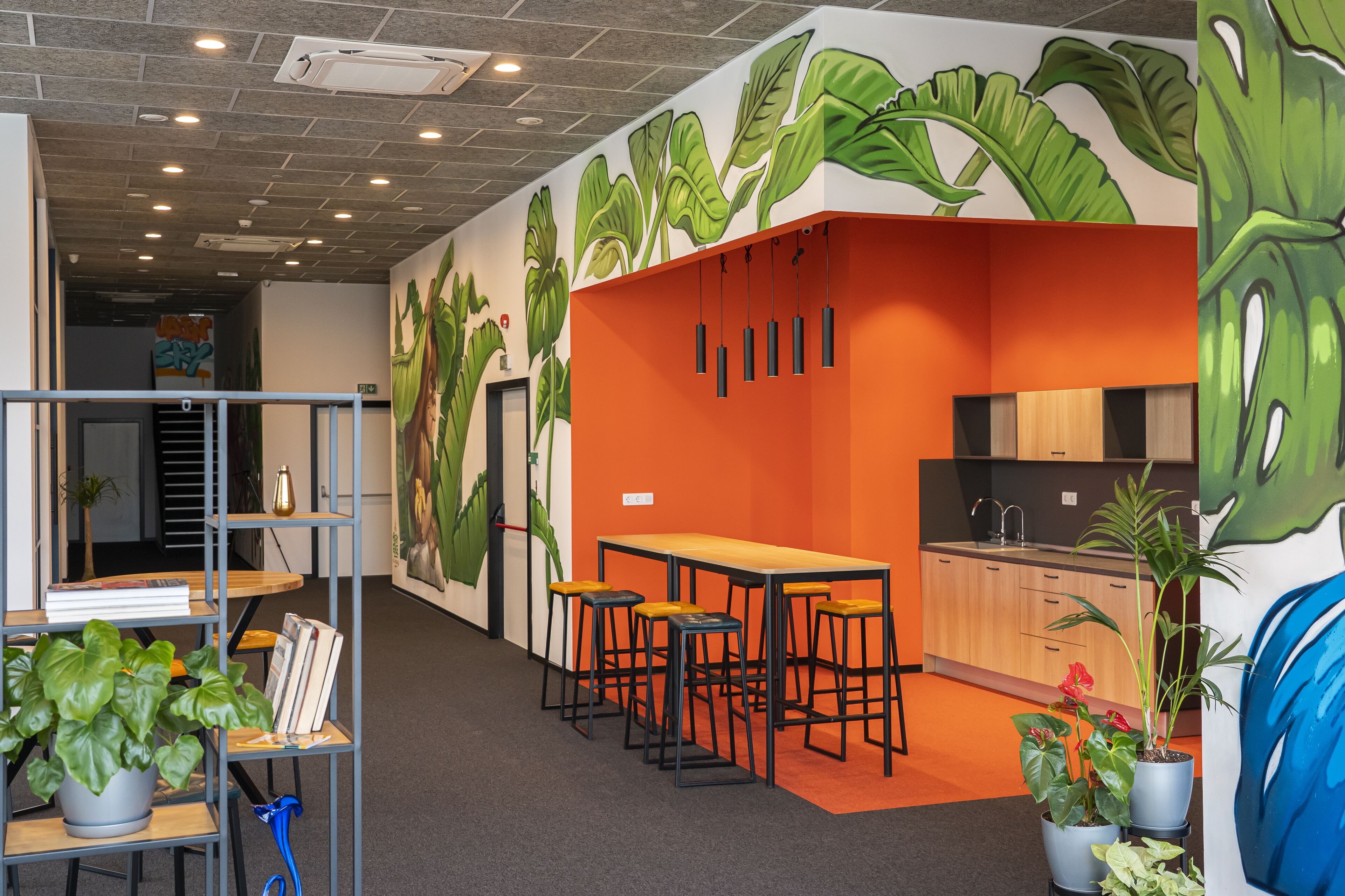 A modern office space with a vibrant orange accent wall, a long table with bar stools, and a variety of potted plants.