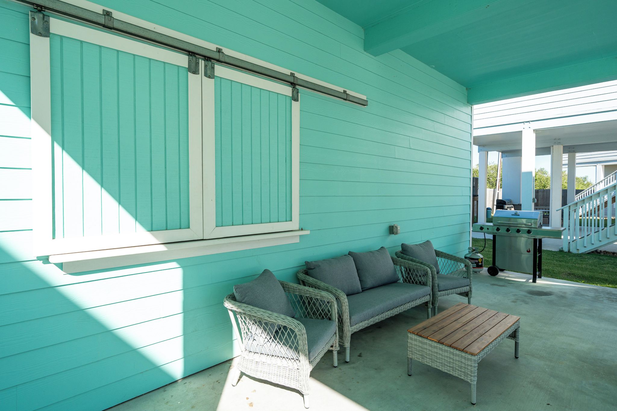 A covered patio area with a turquoise exterior, wicker furniture, and a barbecue grill.