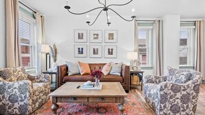 Una sala de estar con un sofá marrón, sillones estampados, una mesa de centro de madera y una araña.
