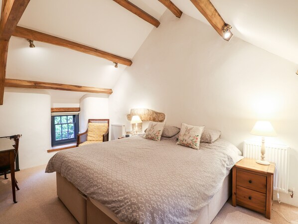A bedroom with a large bed, wooden beams, a window, a chair, and a nightstand with a lamp.
