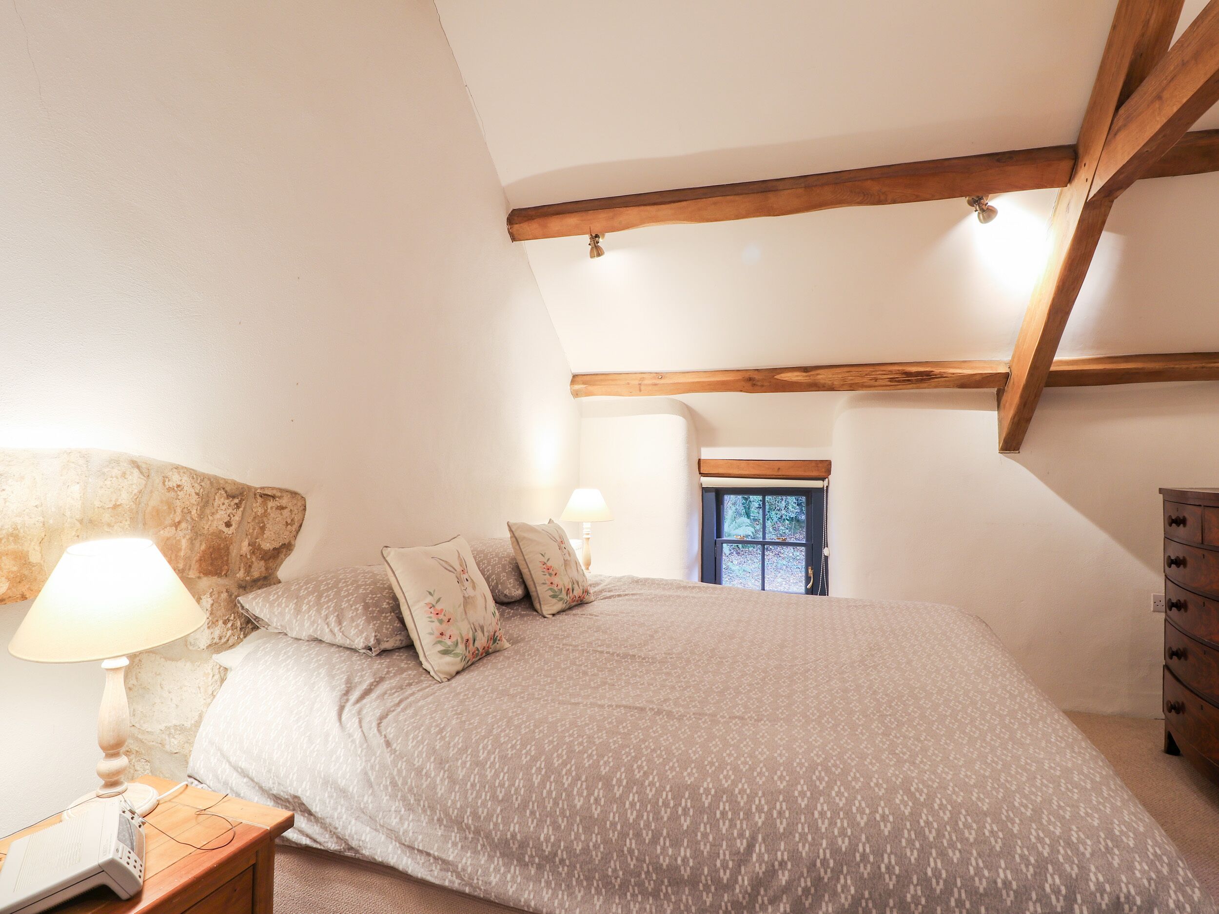 A bedroom with a bed, a nightstand with a lamp, a dresser, and a window with a view of trees.