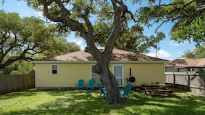A yellow house with a large tree in front, a wooden deck, and three blue chairs. - Coastal Soul by Miss Kitty’s (Aransas Pass)