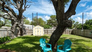 A backyard with a large tree, two turquoise Adirondack chairs, a wooden fence, and a shed. - Coastal Soul by Miss Kitty’s (Aransas Pass)