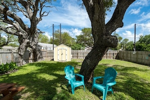 A backyard with a large tree, two turquoise Adirondack chairs, a wooden fence, and a shed. - Coastal Soul by Miss Kitty’s (Aransas Pass)
