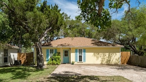 A single-story house with a yellow exterior, a brown roof, and blue shutters. There is a front yard with a paved driveway and a wooden fence. - Coastal Soul by Miss Kitty’s (Aransas Pass)