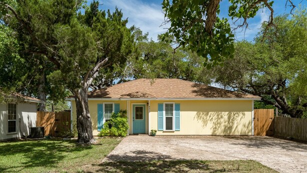 A single-story house with a yellow exterior, a brown roof, and blue shutters. There is a front yard with a paved driveway and a wooden fence. - Coastal Soul by Miss Kitty’s (Aransas Pass)