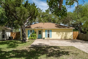 A single-story house with a yellow exterior, a brown roof, and blue shutters. There is a front yard with a paved driveway and a wooden fence. - Coastal Soul by Miss Kitty’s (Aransas Pass)
