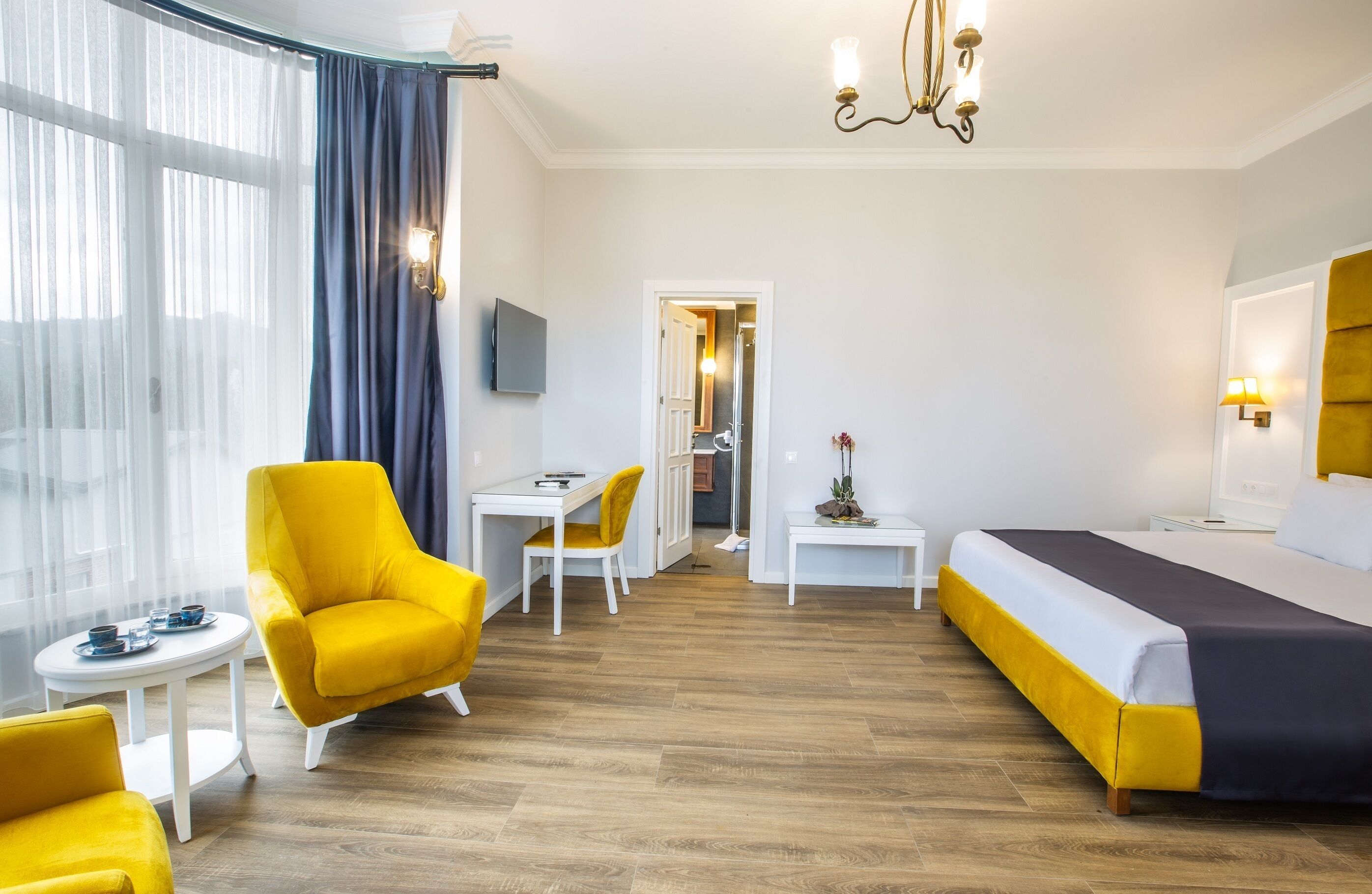 A hotel room with a bed, two yellow armchairs, a small table, a TV, and a view of the hallway.